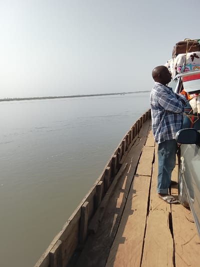 A ferry traverses a wide river in Ibi, Nigeria. On the vessel's wooden deck, a bald, middle-aged man stands with arms crossed, facing left and observing the water. He is dressed in a blue and white plaid long-sleeved shirt, blue jeans, and dark sandals. To his right, the front section of a light-colored vehicle, likely a sedan or SUV, is parked, its roof burdened with various items of luggage, including a brown suitcase and several wrapped bundles. The river's surface is calm, displaying a grey-green tint with subtle ripples, stretching towards a distant, low-lying shoreline. A small, dark boat is visible far in the background on the water. The sky is a uniform pale blue or light grey, suggesting a hazy or overcast day. Shadows cast on the wooden deck indicate sunlight originating from the right.
