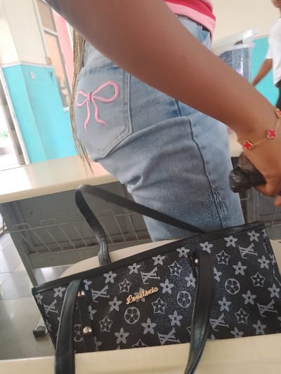 A person in an indoor setting, possibly a classroom in Umueshi, Nigeria, is captured from the waist down. They are wearing light blue jeans with a pink bow design on the back pocket and a red and gold bracelet on their right wrist. A black handbag with light grey flower and "LV" logo designs, and "Lovetonic" in gold lettering, sits on a white table in front of them. A light-colored desk and a blue and pink wall are partially visible in the background. Another person, only partially visible in the upper right of the frame, is wearing a white shirt.