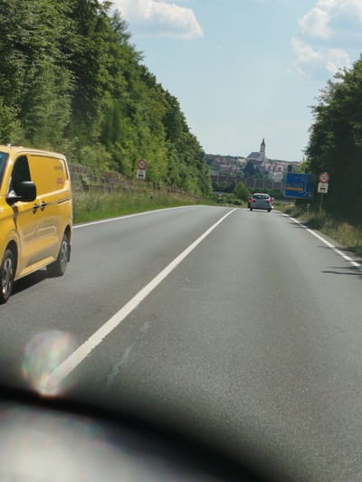 The image captures a road in Kist, Germany, on a clear day. A yellow van is parked on the left side of the road, facing away from the camera. Further down the road, a silver car is driving away from the camera. In the background, a town with a church steeple is visible on a hill. Traffic signs and trees line the sides of the road. The scene appears to be a typical rural or suburban setting.