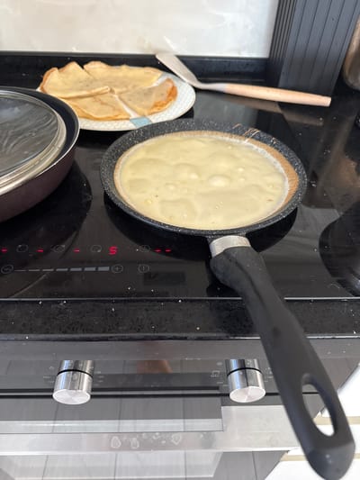 In the city of Uyutnoye, Ukraine, a close-up shot captures a domestic scene focused on cooking. On a black induction stovetop, a non-stick frying pan contains a thin layer of light yellow batter, indicative of crepes or pancakes being prepared. The pan's black handle, featuring a silver-colored metal ferrule, extends towards the viewer. To the left, a brown pot with a transparent lid sits on the stovetop. In the background, a white plate holds four folded crepes, sliced into quarters. A spatula with a light-colored wooden handle rests on the countertop behind the plate. The stovetop displays illuminated red indicators, including the numeral "5," suggesting a cooking setting. Below the stovetop, the glass door of an oven is visible, revealing silver knobs with temperature markings. The overall environment appears to be a kitchen.