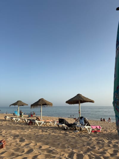 This image depicts a sandy beach under a clear, light blue sky in the late afternoon, identified as a beach near Almancil, Portugal, consistent with Praia da Falésia. The foreground consists of light golden-brown sand exhibiting minor textural variations. Three large, straw-thatched beach umbrellas, each supported by a light-colored pole, are positioned in the mid-ground, extending towards the right side of the frame. In front of and between these umbrellas are multiple white or light-colored sunbeds. One individual is observed lying prone on a sunbed in the central-left area. Other individuals are seated or reclined on adjacent sunbeds, with various items of clothing and towels draped over the furniture. The background features a calm, light blue-green ocean with a distinct horizon line. Multiple individuals are present in the water, either swimming or wading, and additional people are visible along the distant shoreline. A light blue beach tent is positioned on the far left near the water. The extreme right foreground is partially framed by a colorful object displaying an ornate blue and white pattern.
