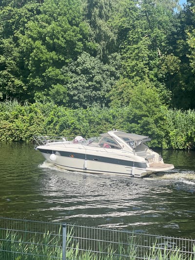 In Teltow, Germany, a white and gray motorboat travels on the Havel River. The boat has a covered cabin and a wooden deck at the stern, with white fenders attached to its sides. It creates a wake as it moves through the water. The riverbanks are lined with dense green trees and reeds. In the foreground, a metal fence and tall green grass are visible. The sky is partly visible through the trees.