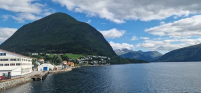 The image shows a scenic view of the city of Volda, Norway, with a focus on its harbor and surrounding landscape. In the foreground, a body of water, likely a fjord, stretches across the scene. To the left, a prominent industrial building with a white facade and multiple windows is visible, adjacent to a stone-covered embankment. A red forklift is parked near the building, and a dock extends into the water.

Beyond the industrial structures, a charming residential area is nestled on the slope of a large, verdant mountain. The houses are a mix of colors, including white, orange, and yellow, with red roofs, creating a picturesque contrast with the lush greenery. The mountain itself is densely covered with trees, with a clear cut on its upper portion that creates a dramatic visual.

In the background, a series of majestic mountains rise, some of which are capped with snow, adding to the dramatic and rugged beauty of the Norwegian landscape. The sky above is a vibrant blue, dotted with fluffy white clouds, suggesting a pleasant day. The overall impression is one of a tranquil and picturesque settlement embraced by the powerful natural beauty of the Norwegian fjords and mountains.