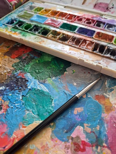 The image displays an overhead, close-up shot of an artist's palette covered in various thick, textured paint splatters. A white plastic watercolor paint set, filled with numerous colorful pans of dried and used paint, rests diagonally across the top half of the frame. A paintbrush with a black handle and silver ferrule lies across the lower portion of the painted palette, angled from the bottom left towards the center right. The background is out of focus and appears to be a patterned fabric. The scene is set on a flat surface, with the lighting suggesting an indoor environment. There is no discernible information about the city of Popovo, Bulgaria, within the visual content of the image.
