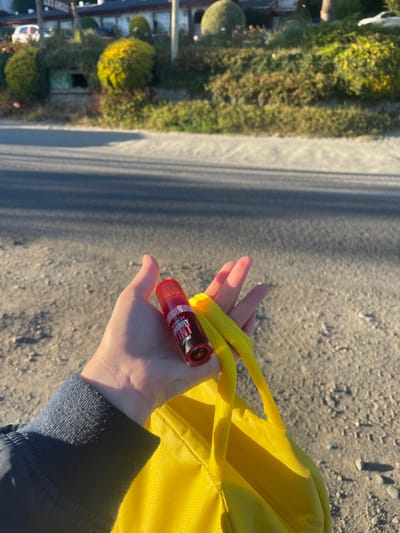 A human left hand, wearing a dark blue long-sleeved garment at the wrist, is centered in the lower portion of the frame. The hand holds a translucent red cylindrical container, labeled "PINK 21 CHERRY LIP TINT," in its palm. The strap of a bright yellow fabric bag is also grasped by the fingers. The immediate ground consists of a dark asphalt road with subtle parallel shadows, transitioning to a lighter, textured surface to the right. The background features a sloping bank of dense green foliage, including several spherical and natural-form bushes. Partially visible behind the foliage is a light-colored building with a dark roofline, displaying a sign reading "LOS ARCOS," indicating the location as San Carlos de Bariloche, Argentina. Two light-colored automobiles are partially visible on the distant left and right sides of the background. Strong directional sunlight casts pronounced shadows across the ground and the hand.
