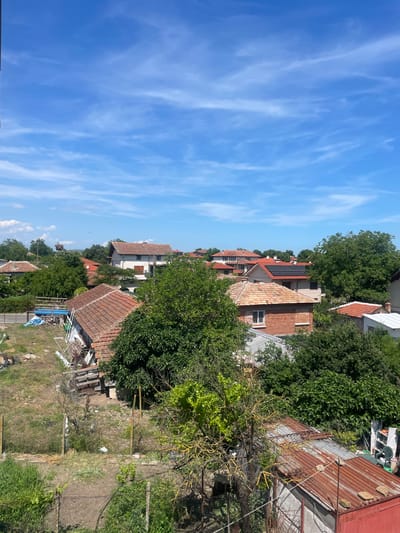 This is an aerial, eye-level, exterior, medium shot of a residential area in Debelt, Bulgaria, on a clear day. The sky is blue with scattered white clouds. The foreground features a cluttered yard with a rusted metal shed, various gardening tools, and overgrown vegetation. Behind the shed, a weathered wooden fence separates the yard from a more manicured area. In the midground, several houses with red tile roofs are visible, surrounded by lush green trees. One house has solar panels on its roof. In the background, more houses and trees are spread across the landscape. A large stork's nest is visible on a utility pole to the left of the frame, with two birds flying in the distance. The scene depicts a tranquil, semi-rural setting.