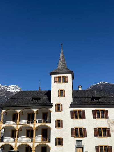The image displays the Landvogteischloss (Bailiff's Castle) located in the city of Visp, Switzerland, under a clear, bright blue sky. The multi-story building features white stucco walls and a dark gray slate roof with dormer windows. A prominent central tower rises from the building, transitioning from a square base to a tall, slender conical spire covered in dark wooden shingles, topped with a metallic finial.

Numerous rectangular windows with distinctive yellow/gold frames and dark brown wooden shutters are visible across the facade. Some shutters are open, others are closed. On the left side of the building, a multi-tiered arched loggia or balcony structure is present, supported by yellow/gold columns and featuring dark metal railings. A smaller spire topped with a cross is visible on a roof section behind the main tower, towards the left. On the right side of the main building, a sundial is affixed to the wall, displaying Roman numerals.

In the background, snow-capped mountains are visible on both the left and right sides of the building, indicating an alpine environment. No individuals or active interactions are depicted within the scene. The lighting suggests direct sunlight, casting subtle shadows on the building's facade.