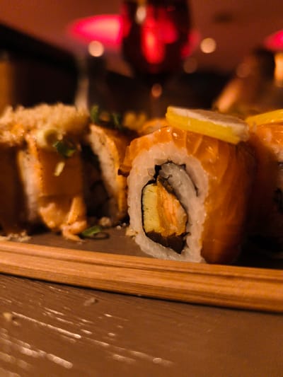 This is a close-up, low-angle shot of a plate of sushi. The sushi rolls are arranged horizontally in the frame, with chopsticks resting in front of them. The main subject is the sushi, which appears to be made with salmon and other ingredients, topped with thin slices of lemon. The lighting is warm and atmospheric, suggesting an indoor setting, possibly a restaurant, at night. The background is blurred, with soft red and pink lights creating a bokeh effect, hinting at ambient lighting or decorations. The surface beneath the sushi and chopsticks is a warm, brown tone, possibly a table or serving platter. The focus is sharp on the sushi in the foreground, particularly the roll with the lemon slice, revealing the textures of the rice, nori, salmon, and other fillings. The overall impression is one of a delicious meal, presented in an inviting and intimate setting. No people or text are visible.