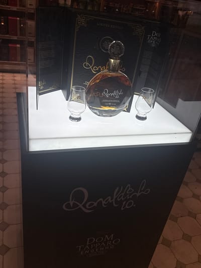 The scene depicts a product display in a retail environment in Guarulhos, Brazil. A dark-colored display stand, branded with "Ronaldinho 10." in white cursive script and "DOM TAPPARO ENGENHO Desde 1978," supports a brightly illuminated white platform. On this platform, a clear, round bottle of "Ronaldinho 10. CACHAÇA EXTRA PREMIUM AGED 10 YEARS 700 ml 40% Alc./Vol." containing an amber liquid is prominently displayed. The bottle features a faceted clear stopper. Flanking the bottle are two empty clear tasting glasses, each adorned with a gold geometric pattern near the rim. Behind the bottle, an open, black, gold-accented presentation box, labeled "LIMITED EDITION" and "DOM TAPPARO ENGENHO Desde 1978," stands upright. The interior of the box reveals partially legible text, including mentions of Ronaldinho. The background, visible through a glass partition, shows dimly lit shelves stocked with various bottles, characteristic of a liquor store or duty-free shop. The floor in the foreground features a patterned tile design with repeating hexagonal and square shapes in light and dark brown tones. No individuals are present in the image.