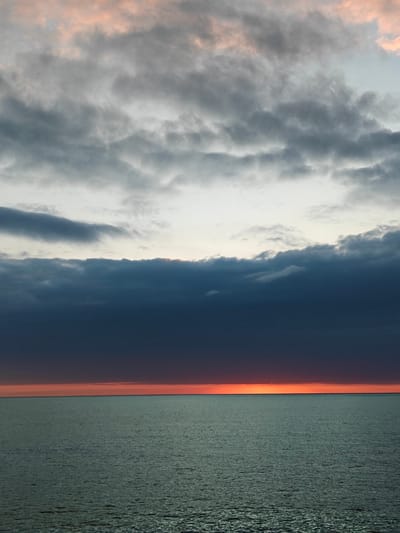 This vertical shot captures a dramatic sunset or sunrise over a vast body of water, likely the Black Sea near Kobuleti, Georgia. The scene is dominated by a layered sky and the calm sea surface meeting at a sharp horizon line.

The upper part of the sky features scattered, textured grey clouds, with soft hints of pale pink and orange light peeking through, suggesting the last or first rays of the sun. Below these, a thick, dense band of dark, heavy storm clouds stretches horizontally across the mid-sky, creating a stark, almost ominous barrier.

Below this dark cloud layer, a brilliant, intense strip of orange and red light glows along the horizon line, where the sky meets the water. This vibrant band indicates the position of the sun, which is obscured by the clouds above but still casts its powerful color.

The lower half of the image is filled by the calm, greenish-blue surface of the sea, which appears vast and slightly textured with ripples from distant waves. The water reflects the muted light from the sky, creating subtle variations in tone.

The overall impression is one of natural grandeur and tranquility, with the dramatic interplay of light and shadow, and warm and cool tones, defining the mood of the changing day. There is no visible text in the image.