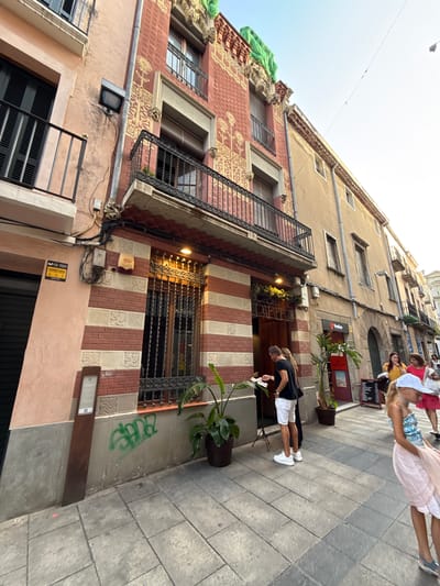 This is a ground-level, slightly upward-angled, full shot image of a street scene in Mataró, Spain. The foreground is dominated by a paved sidewalk made of grey square tiles, with a brown, rectangular information stand positioned on the left. Green graffiti is visible on the lower portion of a building wall to the right of the stand.

The central focus is a multi-story building with a facade featuring red brick bands interspersed with cream-colored decorative panels. The lower part of the building has a large window with ornate black metal bars. Above this, a balcony with similar black metal railings extends across the facade. The building also has yellow patterned embellishments on its upper sections, and a green tarpaulin is draped over the very top. The building's entrance, marked with a sign reading "CASA PARERA," is partially obscured by two people.

To the left of the main building is another building with a light pink facade, featuring a black-shuttered window and a black metal balcony. Electrical conduits and junction boxes are visible on the pink and red facades.

On the sidewalk to the right of the main building, several people are present. A man in a black t-shirt and white shorts stands near the entrance, looking at a menu. Further down the street, a woman in a yellow top and a woman in a pink top are walking. A young girl in a blue and white top and a pink skirt is also visible, with her back mostly to the camera. There are potted plants on the sidewalk, including a large one in a dark pot in front of the "CASA PARERA" entrance. A red sign with white text is mounted on the adjacent building. The sky is clear and bright.