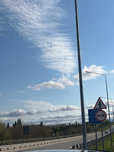 The image presents a view of a road in Yugo-Kamskiy, Russia. Dominating the scene is a light blue sky streaked with wispy cirrus clouds. Adjacent to the road, a dense line of trees borders the pathway. Road signs, including a triangular hazard sign and a circular sign with teacups, are visible on the roadside. A modern lamppost is also present. A section of the road itself can be seen, as well as the barrier separating the road from the side.