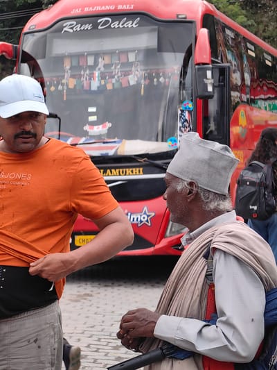 The image shows a street scene in Siyal, India. Two men are in the foreground, standing near a red bus. The man on the left is wearing an orange t-shirt and a white baseball cap, and is pulling at the hem of his shirt. The man on the right is wearing a grey cap and a light-colored shirt, with a tan shawl draped around his shoulders. He appears to be sitting or leaning, possibly in a wheelchair, and is holding something in his hands. The bus behind them has writing on it that includes "JAI BAJRANG BALI" and "Ram Dalal". Small decorative flags are hanging from the bus's interior. The ground appears to be paved with cobblestones or similar material, and some trees are visible in the background. A backpack is visible on another person further back in the image.