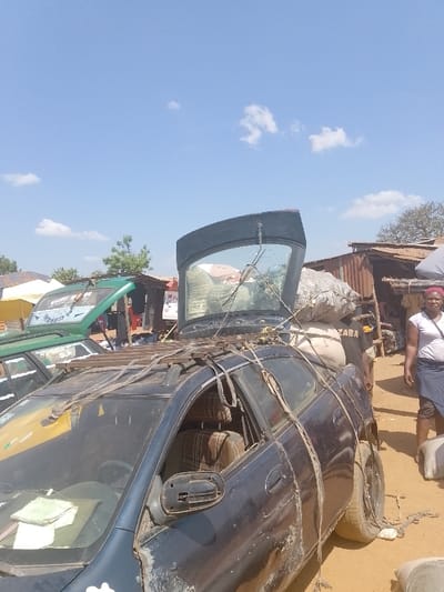 This image depicts an outdoor market or roadside scene in Omupo, Nigeria, under a clear blue sky with scattered cumulus clouds. The foreground and midground feature a heavily modified, dark-colored sedan (possibly black or dark blue) positioned on a dirt ground. The vehicle's rear hatch is open, revealing a significant load of large, light-colored sacks filled with goods, which are piled high and secured with visible rope or fabric straps. A rudimentary metal roof rack is affixed to the car's roof, from which additional straps extend down the sides, wrapping around the vehicle's body. The car displays considerable wear, including dirt accumulation, paint degradation, and a damaged or incomplete driver's side mirror. A green, rectangular object rests on the dashboard. To the left of this sedan, a portion of a green van or bus with an open hood is visible.
To the right of the dark sedan, a woman of dark complexion stands facing the viewer. She is attired in a light-colored (likely white) short-sleeved top, a dark-colored skirt, and a red headwrap. Her arms are positioned at her sides. Partially obscured behind the dark sedan and to the left of the woman, a man's torso and right arm are visible. He is wearing a dark t-shirt with the word "ZARA" partially legible and appears to be in proximity to or handling additional sacks of goods.
The background includes several simple structures with corrugated metal roofs and walls, indicative of market stalls or small shops. One such structure directly behind the dark sedan features a partially visible sign beginning with the letter "P." Scattered trees and bushes are also present in the background.