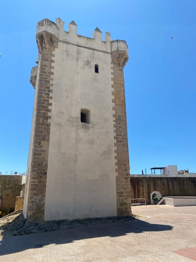 The image captures a square, whitewashed tower with stone battlements and rectangular cutouts against a clear blue sky. The tower is located in the city of Conil de la Frontera, Spain, specifically in the Torre de Guzmán. The tower is made of stone and whitewashed plaster, with distinct crenellations along its upper edge. Two small, rectangular windows are visible on the main face of the tower. The base of the tower is surrounded by a cobblestone ground and some rubble. In the background, a low, dark stone wall stretches across the scene, topped with modern buildings and structures. A small, arched entrance with steps is visible in the wall. The lighting suggests it is daytime, with the sun casting a long shadow from the tower onto the foreground. No people are clearly visible.