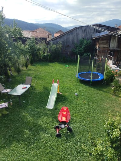 The image shows a backyard with a trampoline, a slide, a child's tricycle, a glass-top table with two chairs, and a few balls on the grass. In the background, there are some wooden sheds and houses with tiled roofs, surrounded by trees and a hill with more houses in the distance. The sky is cloudy. The scene appears to be in a rural or semi-rural area, likely in Rakitovo, Bulgaria, given the architectural style of the buildings and the mountainous backdrop. There are no people visible in the image.