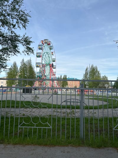 The image depicts a Ferris wheel located in a park in Uray, Russia. The view is partially obstructed by a gray metal fence in the foreground. Behind the fence, there is grass, a paved walkway, trees, and buildings. The Ferris wheel is visible on the left side of the image, towering above the trees. The sky is blue with some clouds.
