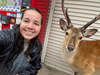 A woman and a male deer with antlers are captured in a selfie-style photograph in Nara, Japan. The woman, positioned on the left, smiles directly at the camera. Her dark hair is damp, and she wears a black rain jacket covered in water droplets. To her right, a Sika deer faces the camera, its reddish-brown fur visible, with prominent dark antlers. In the background behind the woman, a red Japanese vending machine offers various canned beverages, including "Red Bull" and "Lemon" drinks, with prices displayed in Yen. Payment options like Alipay+ are visible. To the right of the vending machine and behind the deer, a light-colored, horizontally corrugated metal roll-down shutter is visible. The ground appears to be wet concrete or asphalt.