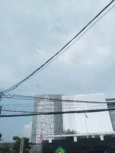 The image captures an exterior view of a building under a light blue, cloudy sky. Numerous black electrical wires run across the upper and middle sections of the frame, obscuring parts of the sky and the building.
In the foreground, at the bottom left, there's a dark metal fence with green foliage partially visible behind it. A red and white circular sign, commonly indicating "no parking" or "no stopping," is partially visible near the fence, with the red diagonal line crossing a white circle.
The main subject of the image is a large, modern building. The building features several distinct sections. On the left side, there's a tall, rectangular section made of gray material, possibly stone or concrete, adorned with large white Arabic calligraphy in a geometric Kufic style. The calligraphy appears to be a verse from the Quran, "Iqra' bismi rabbika alladzi khalaq" (Read in the name of your Lord who created). To the right of this gray section is a taller part of the building with a reflective, perhaps glass or white paneling, facade. In front of this reflective section, an Indonesian flag, red and white, is visible, hanging from a flagpole. Further to the right, a darker section of the building with an intricate, geometric pattern is partially visible.
At the very bottom of the image, below the building facade and behind the fence, a dark green sign or barrier is visible. On this sign, white text is discernable, reading "KEMENTERIAN AGAMA" (Ministry of Religious Affairs) and "PUSAT LITERAS" (Literacy Center), indicating the building's function. A green emblem with a star and crescent is also visible on this sign, characteristic of the Indonesian Ministry of Religious Affairs logo.
The overall scene suggests daytime, likely a bright but overcast day due to the diffuse light and visible clouds. The presence of the Indonesian flag and the text in Indonesian and Arabic points to a location in Indonesia.
