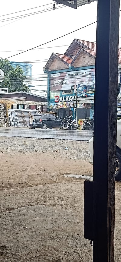 A street scene in Uteungeulinggang, Indonesia, features a dark-colored sedan parked on a wet asphalt road. To the left of the sedan, a person in light-colored clothing stands near a stack of white framed structures, possibly windows or doors. Several motorcycles are parked behind the sedan. A building with a red-tiled roof and blue and white signage stands prominently in the background. The signage displays text in Indonesian, including "ALKAY" and "PRINT KAIN TEXTILE." The overall environment appears to be a commercial area with overcast skies, as evidenced by the wet road and diffused light. A wooden post is in the foreground on the right side of the image.