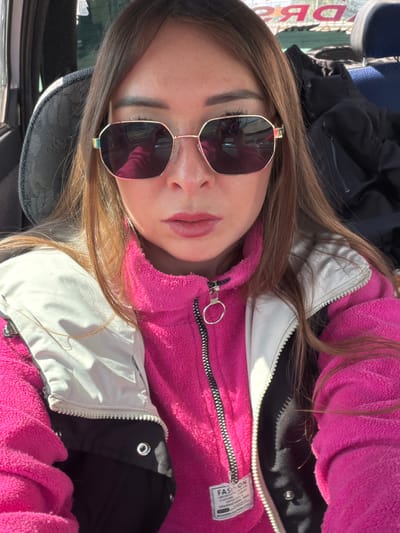 A medium shot captures a woman from the chest up, seated inside a vehicle. She is wearing oversized hexagonal sunglasses with gold frames, a bright pink fleece jacket, and a white and black puffer vest. Her hair is long and brown, falling around her shoulders. The background shows a blurred image of a sign with Cyrillic lettering, possibly indicating a location in Yarykovo, Russia, and the interior of the vehicle. The lighting suggests it is daytime.