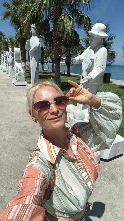 A blonde woman with sunglasses poses for a selfie outdoors. She is wearing a striped shirt and her right hand is raised to adjust her sunglasses. Behind her, there are palm trees, white statues lined up, and the sea with a cruise ship in the distance. The scene appears to be in a public area, possibly a promenade or park, given the presence of statues and the waterfront. The architecture of the statues suggests a modern art installation. The environment is sunny and clear, indicating a warm day. The location is Kuşadası, Turkey, known for its coastal beauty and tourist attractions.