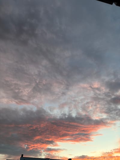 A low-angle, medium shot captures a dramatic sky above Solna, Sweden. The sky is filled with thick, layered clouds. In the upper portion of the frame, the clouds are a dark, stormy grey. As the eye moves down, patches of pink and orange light begin to emerge, suggesting either the light of a setting or rising sun. The bottom of the image features a sliver of the tops of buildings, likely apartment buildings, indicating an urban or suburban setting. The overall mood is one of impending weather or the beauty of a transitional sky.