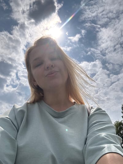 In the city of Breytovo, Russia, a young woman with blonde hair is captured in a selfie outdoors. She is wearing a light blue sweatshirt and looking towards the camera with a slight smile. The background is dominated by a bright, cloudy sky with the sun shining prominently, creating lens flare and a subtle rainbow effect. The clouds are scattered and appear to be in motion, suggesting a windy day. The woman's hair is also being blown by the wind. The image is taken from a low angle, emphasizing the sky and the woman's face. A small portion of a tree can be seen on the right side of the frame.