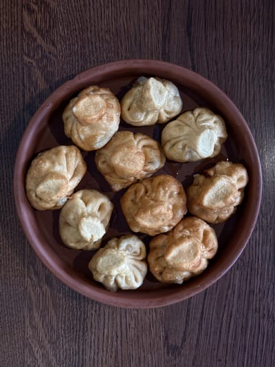 A top-down shot depicts a brown clay plate filled with approximately ten beige and light brown dumplings. The dumplings are irregularly shaped, with crimped edges. The plate rests on a dark brown, textured wooden surface. The scene appears to be indoors. There is no evidence suggesting this photo was taken in the city of Kurchatov, Russia.