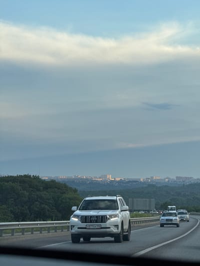 This media file depicts a view from a vehicle traveling on a highway. The primary subjects are several cars on the road, including a white SUV in the foreground with a license plate that reads "K877AA 126". The scene also includes a city skyline in the distance, suggesting an urban or semi-urban setting. Trees line the side of the highway, and the sky is filled with clouds, indicating overcast weather. The time of day appears to be late afternoon or early evening, given the soft, diffused light. The overall mood is neutral, focusing on the transportation aspect of the scene. The location context provided is Novomar'evskaya, Russia.