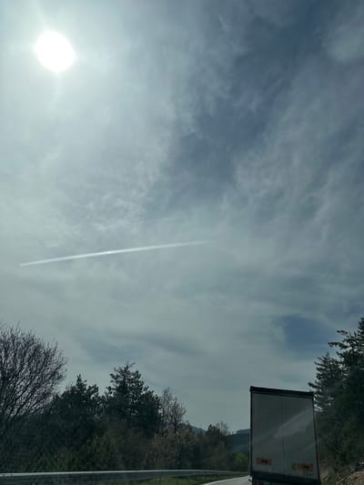 The image captures a bright, partly cloudy daytime sky with natural and man-made elements. The sun is prominent on the left side, appearing as a brilliant white orb, partially obscured by light clouds. A distinct, straight white contrail from an aircraft extends diagonally across the sky from the upper right, passing near the sun's glare.
The sky is filled with wispy, stretched-out clouds, ranging from thin, translucent layers to denser, more defined formations, primarily in shades of blue and grey. On the right side of the frame, a line of trees forms a dense boundary. Some trees appear to be deciduous with bare branches, suggesting late winter or early spring, while others retain their foliage.
Partially visible on the far right is the rear of a large white semi-trailer truck. Small, indistinct markings or logos are present on its back. Below the truck and trees, a metal road barrier runs horizontally, indicating the presence of a highway or main road. In the distant background, beyond the trees and truck, faint outlines of hills or mountains are discernible.
The scene depicts a typical view from a road in a rural or semi-rural landscape, possibly in Dobrinovo, Bulgaria, as indicated by the location context. There is no specific activity or event taking place, rather it is a snapshot of the environment under a bright, cloud-streaked sky. No legible text is visible other than the faint, unidentifiable markings on the truck.