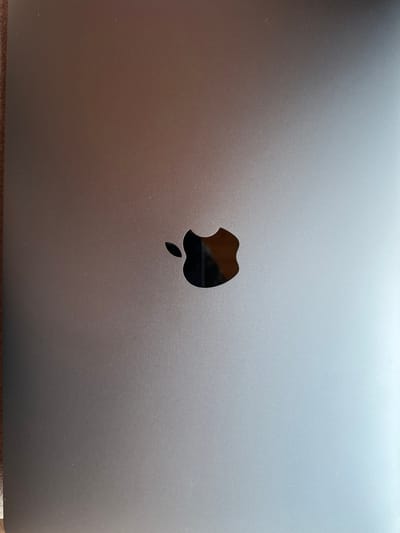 A close-up, overhead shot shows the Apple logo on a laptop. The logo is dark, appearing black or a very deep gray, and is centered on the laptop's metallic casing, which is a muted silver color with a brushed texture. The lighting creates a subtle gradient across the laptop surface, brighter on the right side and darker on the left. The background consists of a brown surface, possibly a table, with a visible wood grain texture on the left edge of the image. There are no people visible, and no context is provided regarding the laptop's location in the city of Samshvilde, Georgia.