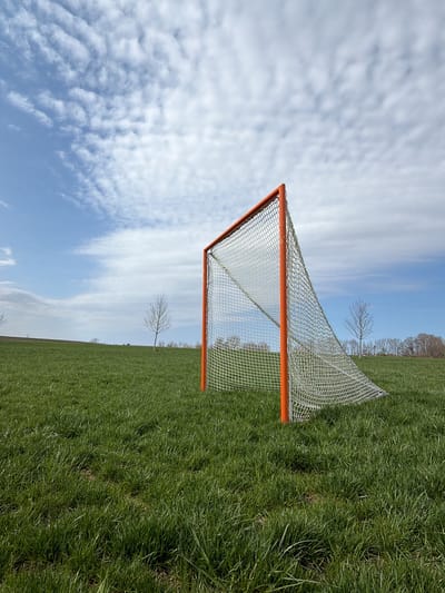 An outdoor, eye-level shot captures a lacrosse goal with a vibrant orange frame and a white net, positioned on a large, open grassy field. The goal is placed slightly to the right of the frame's center, extending into the mid-ground. The foreground and mid-ground are covered in lush, green grass, which appears somewhat wild and untamed, suggesting a natural or undeveloped sports field.
In the background, the field gently slopes upwards, leading to a horizon dotted with a few bare, slender trees, indicating early spring or late autumn. Beyond these isolated trees, a denser line of deciduous trees forms a subtle, darker band against the sky. The sky itself is a bright blue, filled with numerous white, rippled clouds (likely altocumulus), suggesting fair weather during the daytime. No specific activity or event is taking place, and the scene is devoid of people. There is no visible text in the image. The setting is an open, tranquil field located in West Henrietta, United States.