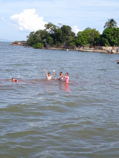 Four children are wading in a body of water. The first child, wearing red, is partially submerged on the left. The next child has their hands raised. Two other children in pink are next to them. Behind them is a shoreline with lush green trees and a few scattered buildings. The sky is partly cloudy. The water is wavy with small ripples.