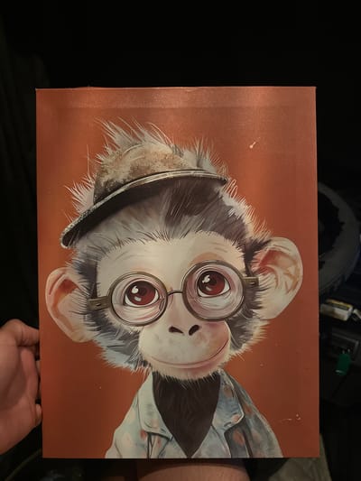 The image is a close-up, front-facing photograph of a painting of a monkey wearing a hat and glasses. The monkey has large, expressive eyes, a small nose, and a slight smile. It is wearing a light-colored shirt with a pattern of pinkish circles. The background of the painting is a solid orange-brown color. A human hand is visible on the left side of the image, holding the painting. The setting appears to be indoors, with a dark background suggesting a room. There is no discernible information that would place this image in Cabanillas del Campo, Spain.