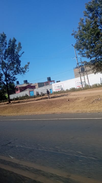 The image captures an outdoor scene, likely taken from a moving vehicle, offering a view of a roadside area under a clear, bright blue sky.
In the foreground, the lower half of the image is dominated by a paved road, dark gray or black in color, with a faint lighter line visible along its edge. The road surface shows some texture and hints of dust or dirt. Above the road, there's a wide, unpaved dirt shoulder or embankment, colored in shades of brownish-orange.
In the mid-ground, a white wall or fence runs horizontally across the frame. Behind this wall, several buildings are visible. Some have reddish-brown roofs, and multiple black water tanks are prominently placed on their rooftops. There are also what appear to be blue doors or gates set into the lower part of the wall. To the far right, behind the main white wall, a taller, unfinished concrete structure can be seen.
A single person is walking on the dirt shoulder in the mid-ground, moving from left to right. They are wearing a dark t-shirt and dark pants. Their posture suggests they are looking down or straight ahead.
Along the white wall, at least two identical advertisements are visible. The main text on these signs reads "SANLINK" in large, stylized letters. Below this, smaller text states "Motorcycle parts & accessories" and "Engine Expert". An illustration, possibly of a motorcycle or an engine part, is also part of the logo. The signs use red and black text on a white background.
To the left and right sides of the image, the tops of green-leaved trees are visible, framing the scene. On the right side, a utility pole with power lines strung across the sky is present, extending above the structures.
The weather appears to be sunny and clear, indicative of daytime, likely midday or early afternoon given the high, shadow-less sun on the sky. The overall impression is that of a semi-urban or rural environment alongside a main thoroughfare.