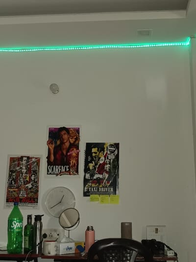 The image shows the interior of a room in Ghaziabad, India, located at 28.682 latitude and 77.41 longitude. The room has a white wall with a green LED light strip mounted on the ceiling. There are three movie posters on the wall: a "Scarface" poster featuring Al Pacino and a woman, a "Taxi Driver" poster featuring Robert De Niro and a woman, and a poster for the movie "Kill Bill" featuring Uma Thurman. There is a clock on the wall, a round mirror on a stand, a bottle of green soda labeled "Sprite" on a table, a silver water bottle, a pink thermos, a brown ceramic mug, a box of facial tissues labeled "Sawan," and a black wicker-backed chair. The room has a desk with a black surface and a black camera on top of it. The camera is positioned in front of the black chair. The room appears to be a bedroom or office space.
