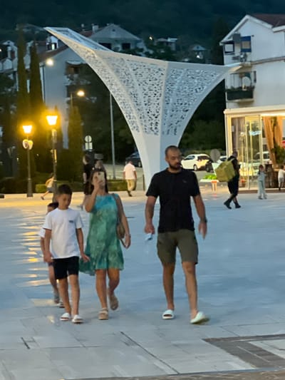 In the city of Kumbor, Montenegro, a group of people are walking on a paved plaza in the evening. A family appears to be walking together, with a man in a black t-shirt and shorts leading, followed by a woman in a blue and green patterned dress holding a phone, and a young boy in a white t-shirt and black shorts. Another child is partially visible behind the boy. In the background, a white modern sculpture with an intricate design is illuminated. The environment includes buildings with white facades, streetlights casting warm yellow glows, trees, and cars parked in the distance. The scene conveys a sense of casual evening activity.