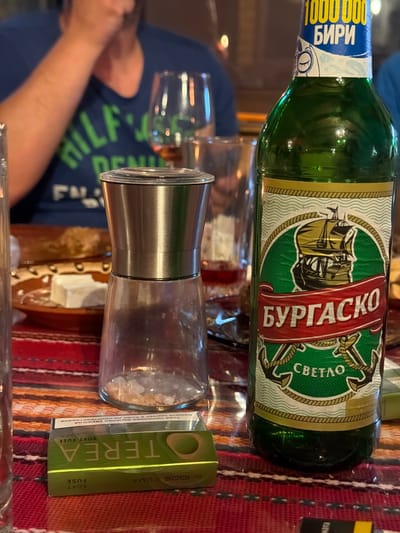The image shows a table setting at what appears to be a restaurant or home in an unidentified location, likely Tsatsarovtsi, Bulgaria. The scene features a bottle of "Burgasko Svetlo" beer, a brand locally produced. Beside it sits a clear glass spice grinder, filled with what looks like coarse salt. Next to the spice grinder is a green package of "TEREA Soft Fuse" for IQOS ILUMA. A partially visible person is seated at the table. The tabletop is covered with a red patterned tablecloth.