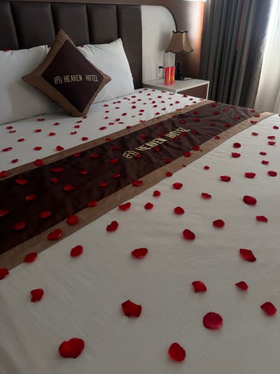 The image shows a bed decorated with red rose petals in a hotel room. The bed has white sheets and a dark brown runner with gold lettering that reads "HEAVEN HOTEL." A brown throw pillow with the same "HEAVEN HOTEL" logo is placed on the left side of the bed. The room has a dark brown headboard, and a nightstand with a lamp and two pink candles is visible on the right. The scene appears to be in a hotel room in Cam Lâm, Vietnam.
