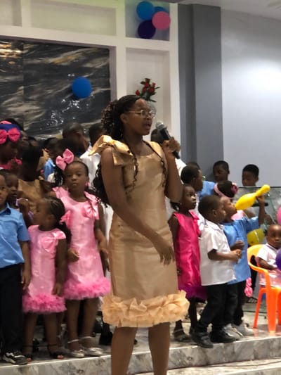 An indoor scene in Gbawe, Ghana, depicts an adult female speaking into a microphone, surrounded by approximately twenty children on a raised platform. The woman, centrally positioned, has dark skin, wears clear-framed glasses, and has long, dark braided hair. She is dressed in a sleeveless, knee-length, champagne-gold dress with ruffled shoulder embellishments and a ruffled hem. Her mouth is open, and she holds a silver microphone in her left hand.

To the left of the woman, several young girls are visible, two prominently dressed in matching bright pink dresses with large pink bows on the bodice and ruffled pink hems. Other children, both boys and girls, stand behind and to the sides of the woman. Their attire includes light blue shirts with dark pants, and white shirts with dark pants. One child on the right holds a yellow, bowling-pin-shaped balloon.

The background features light-colored walls with recessed architectural niches. The upper right niche contains three balloons: one blue, one pink, and one purple. A lower niche holds a small bouquet of red roses. To the upper left, a wall section has a dark, marbled pattern with a single blue balloon attached. The foreground shows light-colored, marble-patterned steps. An orange plastic chair is partially visible on the far right.