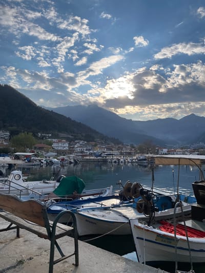 The image depicts a waterfront scene in Skala Potamias, Greece. In the foreground, a concrete pier is lined with several docked boats of varying sizes and types. The boats are close together and some are equipped with fishing gear.

Behind the pier and boats, a town stretches along the shoreline, with buildings featuring white facades and red-tiled roofs. A steep, mountainous landscape rises in the background, partially shrouded in haze. The sky is blue with scattered clouds, and the sun is visible, creating a bright glow.