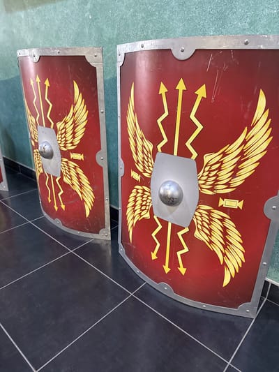 This is a medium shot, eye-level, interior photograph of two Roman scutum shields displayed in a museum or historical exhibition. The shields are identical in design, featuring a red background with a yellow winged emblem, a central silver boss, and yellow lightning bolt motifs. The shields are partially visible, with the left one angled to the right and the right one angled to the left, creating a sense of depth. The shields are framed by metal edging and rivets. The background consists of a mottled green wall and a dark tiled floor with white grout lines. The overall lighting is even and slightly diffused. The location is identified as Sremska Mitrovica, Serbia, specifically within the Srem Museum. There are no people or animals visible in the frame.