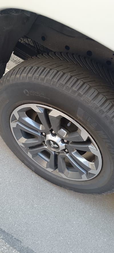 This is a close-up, eye-level shot of a vehicle's tire and wheel. The tire is a Michelin Total Performance tire, as indicated by the visible text on its sidewall. The wheel is dark gray with polished silver accents, featuring a multi-spoke design. At the center of the wheel is a silver emblem with a stylized "X" or star shape, which appears to be the Mitsubishi logo. The tire is mounted on asphalt pavement, suggesting an outdoor setting. The lighting indicates it's daytime, with no specific weather conditions immediately apparent. The image appears to be taken in Schwaikheim, Germany. There are no people or active events visible; it's a still shot focused on the tire and wheel assembly. The overall impression is one of detail and clarity of the vehicle's components.