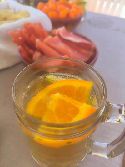The image shows a close-up of a glass of water with orange and lime slices in it. The glass is on a table, which is covered with a light-colored tablecloth. In the background, there are several bowls of food, including what appears to be carrots and possibly some other fruits or vegetables. The scene is outdoors, with a fence visible in the far background. The overall impression is of a refreshing drink served in a casual outdoor setting. The location is identified as Dzērumi, Latvia.