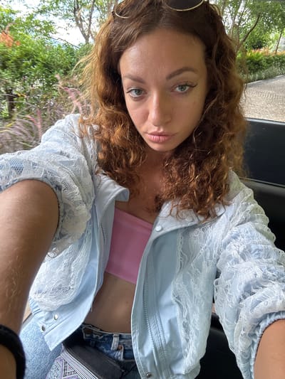 A female individual is depicted in a close-up selfie, seated outdoors in a lush green environment, identified as Mu Cang Chai, Vietnam. She has fair skin, reddish-brown curly hair, light green eyes, and an expression featuring a slight pout. Sunglasses with gold frames are positioned on top of her head. She wears a light blue jacket with intricate white lace detailing overlaying the sleeves and portions of the torso. Beneath the jacket, a pink bandeau top is visible. Her lower body is clad in blue denim jeans, and a black fanny pack with a distinct purple, white, and black geometric pattern on its strap is worn around her waist. Her left arm, showing light hair, is visible in the foreground. The background consists of dense green foliage, including trees and shrubs, with some reddish-purple feathery ornamental grasses in the mid-ground. A textured, possibly cobblestone-paved, path or road is visible on the far right. The overall scene is illuminated by natural daylight.