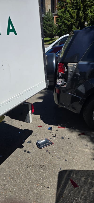 The image shows the aftermath of a vehicle collision. The scene is an asphalt road, likely in a residential area, as indicated by the presence of a building and trees in the background. The primary objects are a dark-colored car with significant damage to its rear, specifically its taillights, and a white trailer or van. Debris from the damaged car, including pieces of red and clear plastic, is scattered across the road.

The car appears to be a sport utility vehicle or a similar model, with its rear bumper and taillight assemblies broken. One taillight is completely detached and lying on the ground, while the other is partially shattered. The white trailer/van is positioned alongside the car, partially obscuring its rear. The side of this vehicle displays green lettering, with the letter "A" clearly visible.

The lighting suggests it is daytime, with harsh shadows cast by the vehicles and trees, indicating a sunny period, likely afternoon. The weather appears clear. There are no people visible in the image. The overall impression is one of a recent accident, with the damaged vehicles and scattered debris serving as evidence of the event. The location context of Sallent de Gállego, Spain, is provided, but there are no specific visual cues within the image that definitively identify this location.