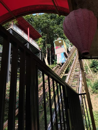 The image depicts an outdoor scene, likely in Gudangkahuripan, Indonesia, featuring a funicular railway ascending a verdant hillside. In the foreground, a dark metal railing provides a partial view of the tracks. The funicular car, a colorful, boxy structure with a yellow cage-like top section, is positioned on the tracks, ascending towards the upper right of the frame. Several individuals are visible inside the car, appearing to be passengers.

Above the funicular, a red awning extends over a building with a similar red roof, suggesting an elevated structure or station. To the right of the funicular, a large, textured pink object, possibly a decorative element or balloon, hangs from an overhead structure. The surrounding environment is characterized by lush green trees and foliage, with hints of flowering plants along the side of the railway. The overall impression is of a tourist attraction or a mode of transport integrated into a natural landscape. The scene is bathed in natural sunlight, indicating it was taken during the daytime.