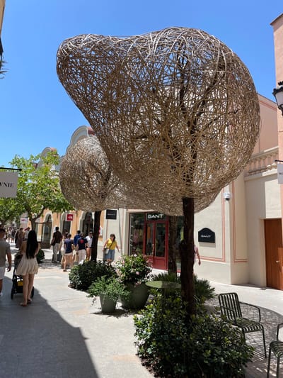 The image depicts an outdoor shopping street in La Roca Village, an outlet shopping destination in La Roca del Vallès, Spain. The scene is illuminated by bright sunlight, casting distinct shadows on the paved walkway. In the foreground, large, abstract tree sculptures, woven from natural fibers, dominate the view. One sculpture is prominently positioned in the center, with another smaller one visible behind it. Lush green plants in large planters are arranged around the base of these sculptures.

Several people are walking along the street. A woman with long dark hair is pushing a stroller, and other shoppers are seen in various groups, some carrying shopping bags. Storefronts line the street, with visible signage for brands like "GANT" and "The North Face." The architecture of the buildings is light-colored with arched windows and decorative trim. The overall atmosphere suggests a pleasant day for shopping.