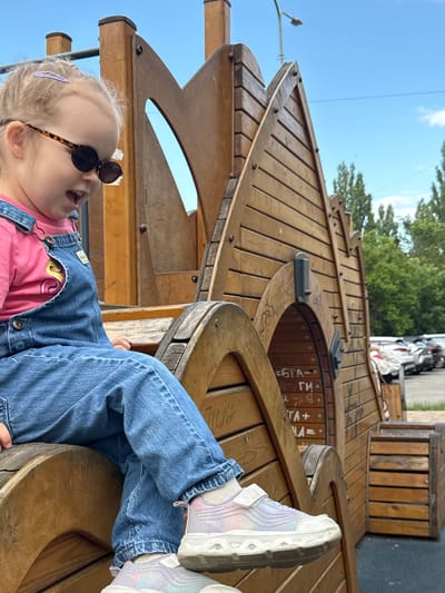 In the image, a young child with blonde hair is wearing denim overalls and a pink shirt. The child also has on leopard-print sunglasses and is looking up with an open mouth, appearing to be in mid-sentence or exclaiming something. The child's legs are propped up on a wooden structure, and they are wearing colorful sneakers.
The setting appears to be a playground made of wood, possibly designed to resemble a ship or a fort, with various wooden planks and curved elements. Behind the playground, trees and a clear blue sky are visible, suggesting it is daytime with good weather. A street and parked cars can be seen in the background, indicating an outdoor urban or suburban environment.
The image is oriented vertically, with the child positioned towards the bottom left. The overall impression is a candid shot of a child enjoying playtime in a unique playground. Some Cyrillic text is visible on the wooden structure, though it is partially obscured and difficult to read completely. Snippets of text like "БРА-", "ГП-", and "ТА+" are discernible. The location context is Верхнерусское, Russia.